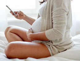 A pregnant woman, seen from neck down, sits on a bed and looks at her phone.