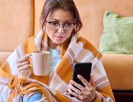 A woman sits on the floor in front of a couch. She's wrapped in a blanket and holding a mug while looking at her phone.