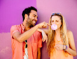 A woman blows a bubble with her gum, and a man beside her playfully pops it.