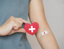 A bandaged arm with a blood drop drawn on the bandage extended as the other hand holds a paper heart with a red cross symbol.